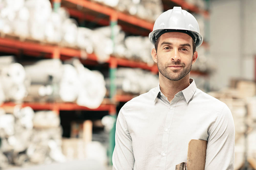 Portrait of a foreman holding a clipboard and wearing a hardhat while standing on a carpet warehouse floor with inventory on shelves in the background