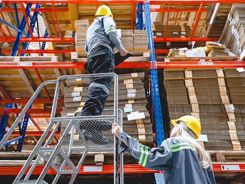 Two factory workers wearing protective helmets and uniforms work together in a cardboard box warehouse. One climbs a safety ladder while arranging paperboard sheets on a high industrial shelf.