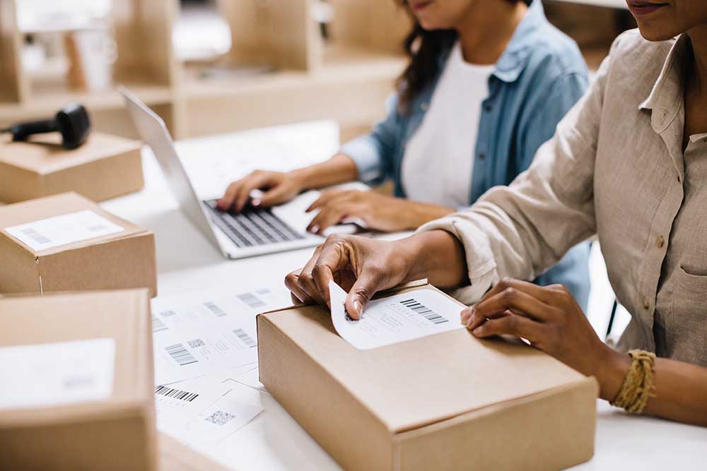 Unrecognizable online store owner sticking a barcode label onto a package box. Businesswoman preparing an order for shipping in a warehouse. Female entrepreneurs running an e-commerce small business.