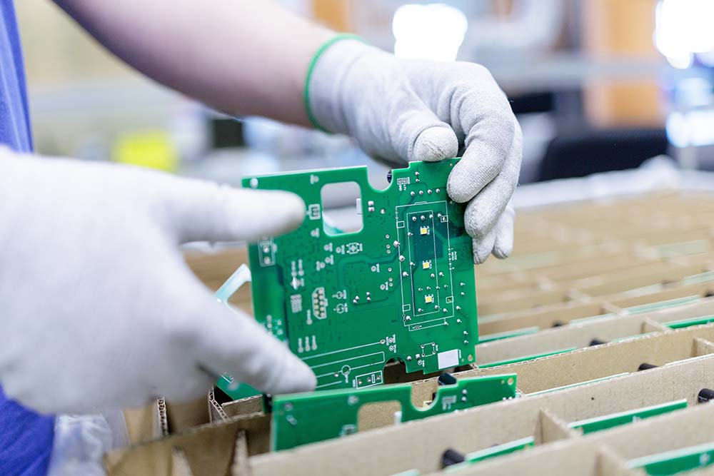 Worker in protective gloves placing green printed circuit board into cardboard tray for safe storage and shipping in electronics plant